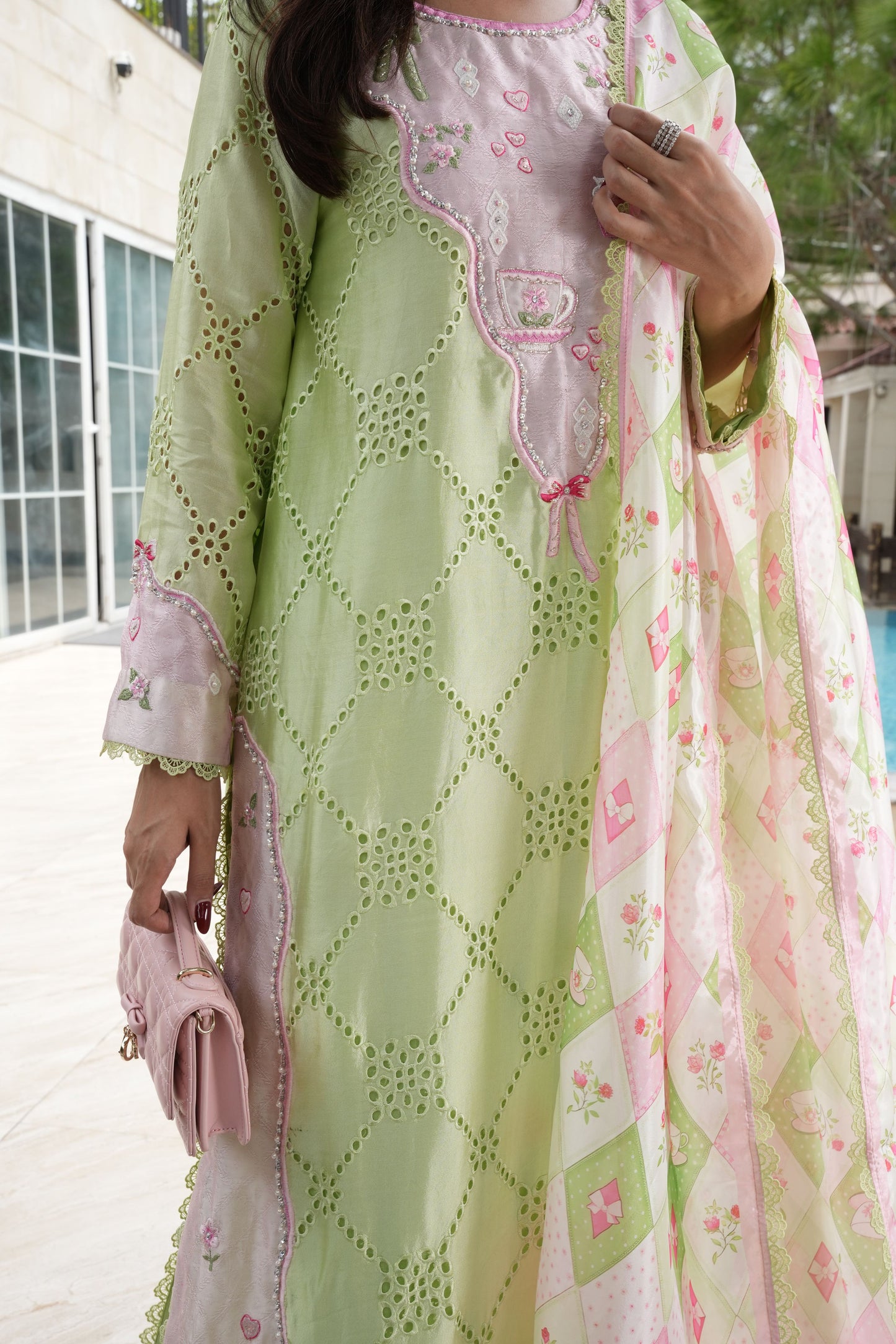 A woman wearing a light green and pink traditional embroidered outfit with a matching dupatta, holding a small pink clutch bag in an outdoor setting.