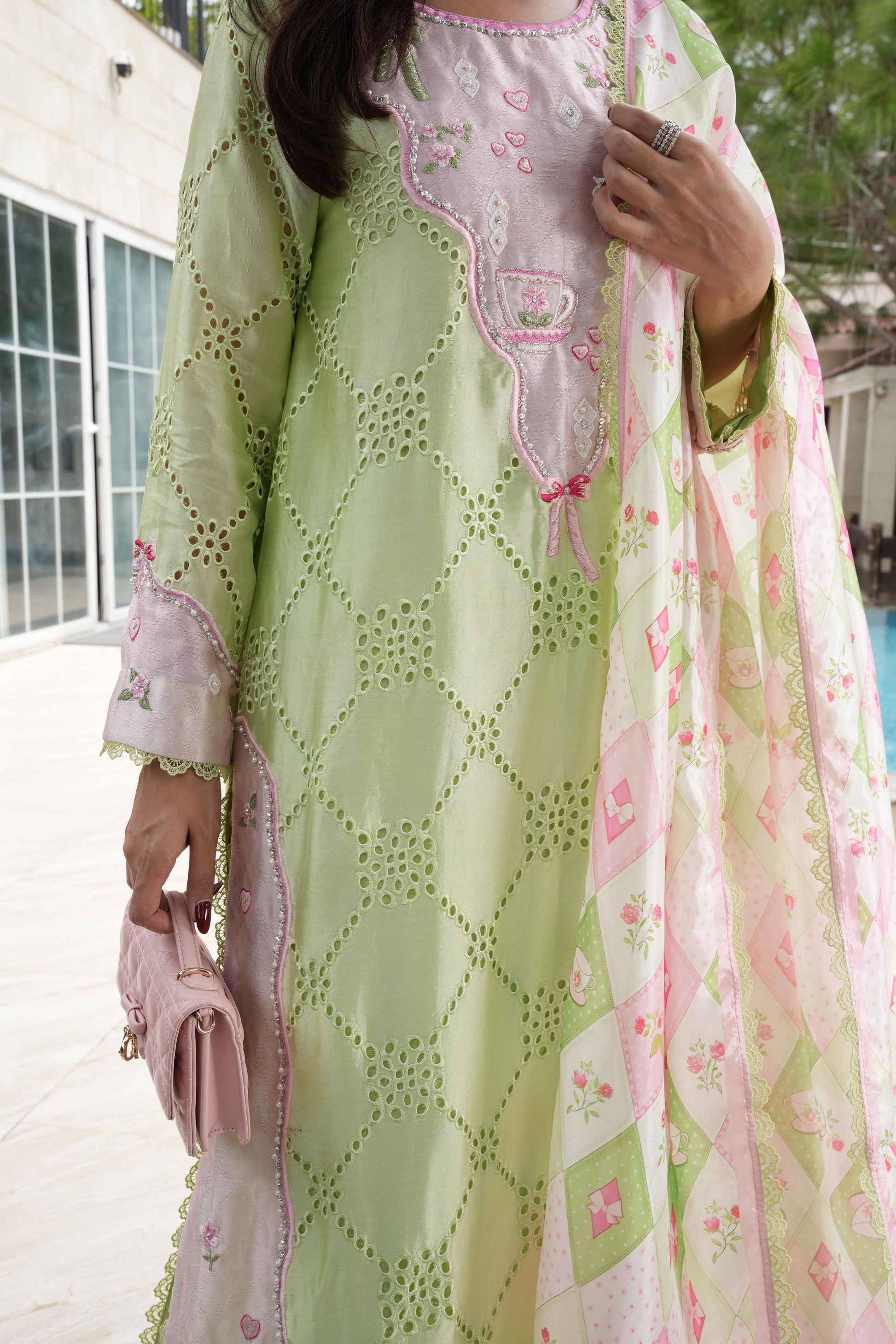 A woman wearing a light green and pink traditional embroidered outfit with a matching dupatta, holding a small pink clutch bag in an outdoor setting.