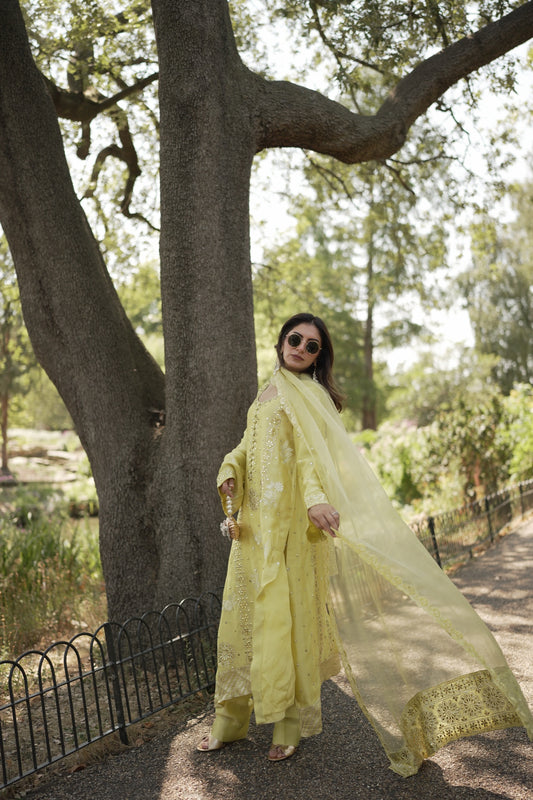 A woman wearing a flowing pale yellow traditional outfit stands outdoors beneath a large tree, holding out the fabric of her dress. The photo is taken from below, with the tree trunk dominating the frame and a path and foliage in the background.